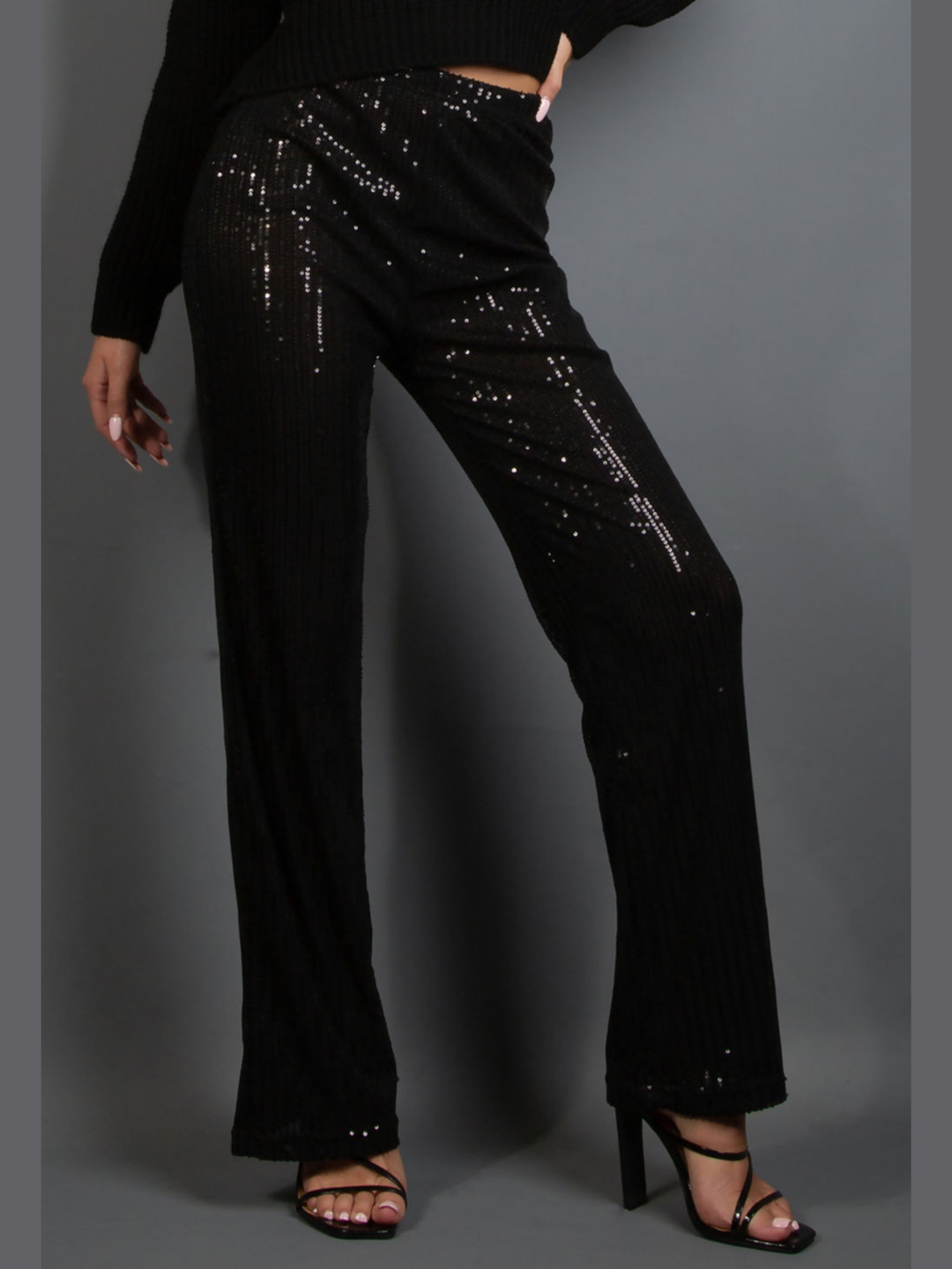Model wears a high waisted black sequin trousers that has a wide leg.  The model faces forward, the front of the wide legged sequinned trousers can be seen. 