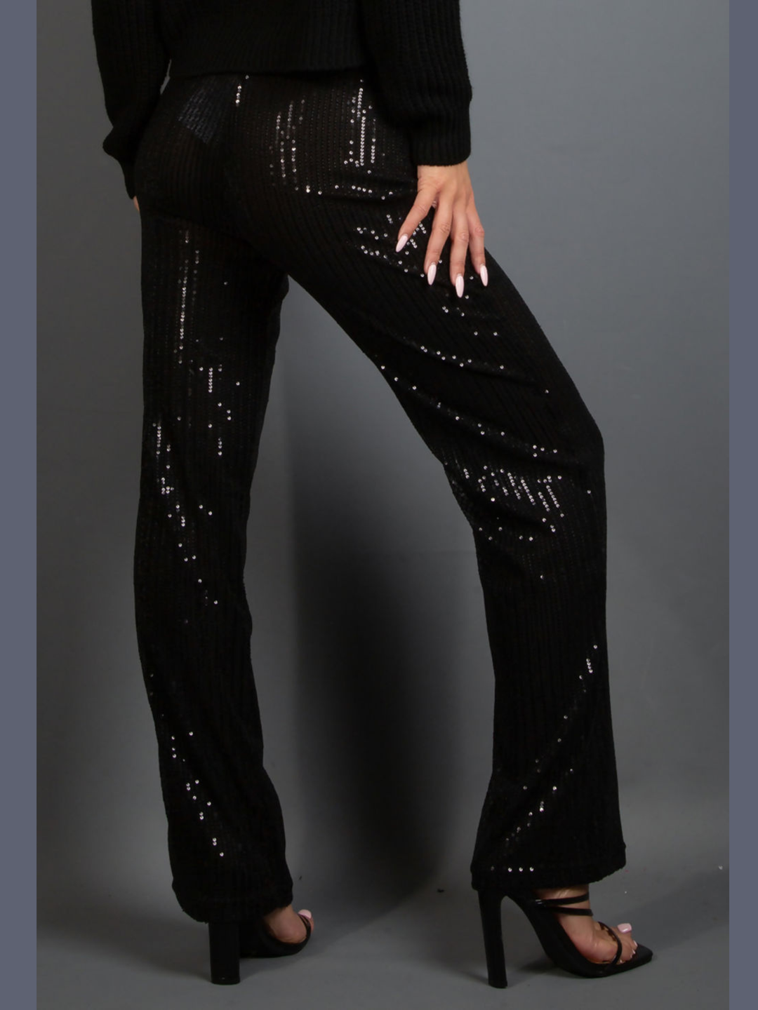 Model wears a high waisted black sequin trousers that has a wide leg.  The model stands with her back to the camera, the back of the black sequinned trousers can be seen. 