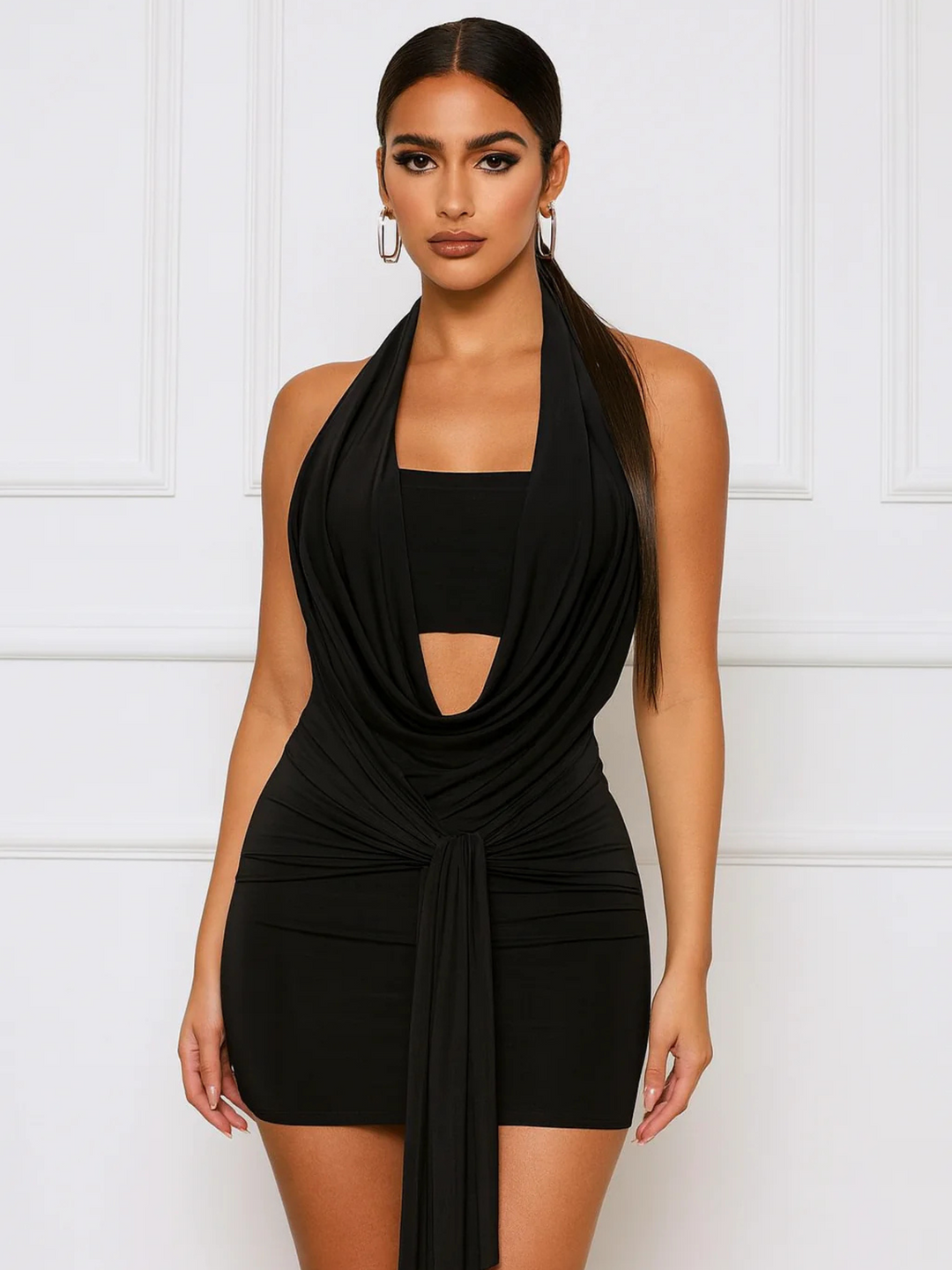Female model stands before a panelled white wall. The model wears slinky black mini dress with a plunging cowl front and a  drape tie front. The model has tanned skin and slicked back long dark hair. 