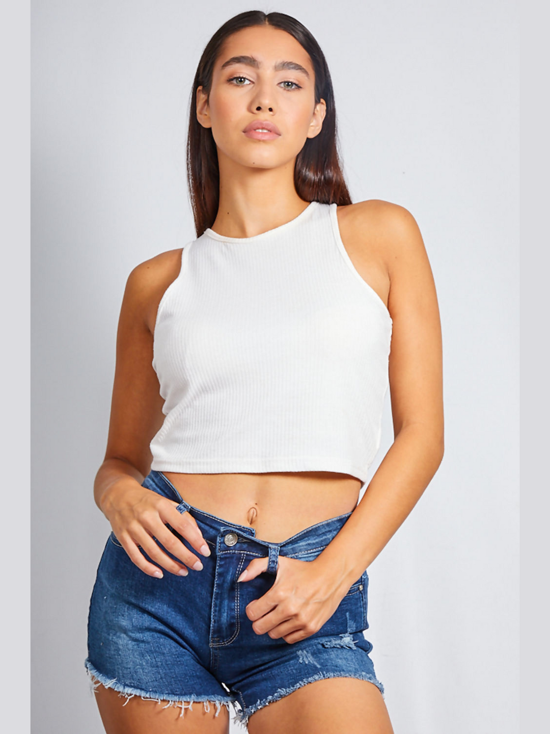 Female model stands in front of a blank grey background, she wears a white ribbed crop top and blue denim shorts with a distressed freyed hem. The model  has tanned skin and long brown hair, she faces the camera and places her fingers in the belt loops of the ditressed denim shorts. 