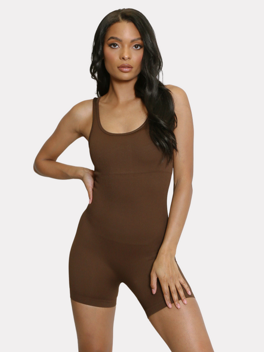 Model wears a brown ribbed unitard jumpsuit with a scoop neck, waist control and open back. Model has light brown skin and long black hair, she stands with one hand on her hip and one hand on her thigh. The model looks directly into the camera. 