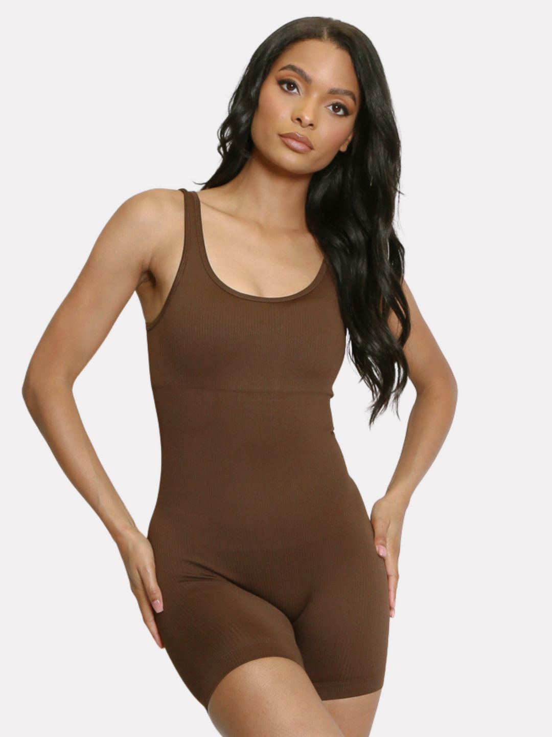 Model wears a brown ribbed unitard jumpsuit with a scoop neck, waist control and open back. Model has light brown skin and long black hair, she stands facing forward and places both hands on her hips, she tilts her head slightly to the side. 