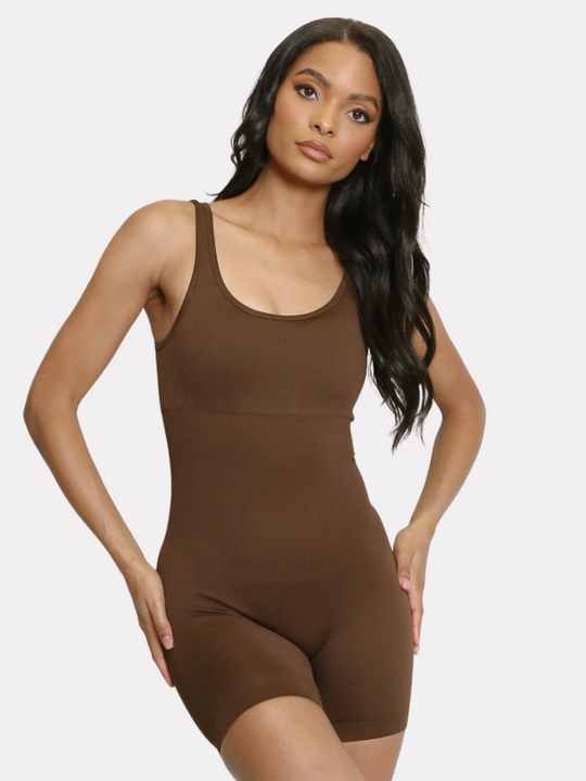 Model wears a brown ribbed unitard jumpsuit with a scoop neck, waist control and open back. Model has light brown skin and long black hair, she stands facing forward and places both hands on her hips, she tilts her head slightly to the side. 