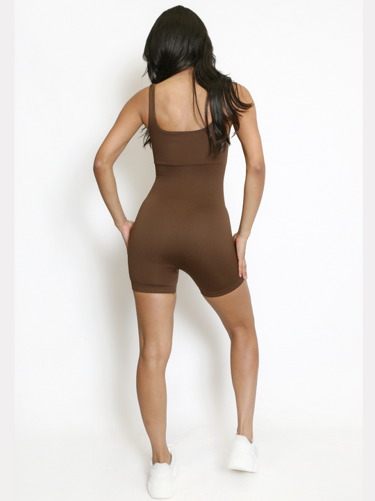 Full length shot. Model wears a brown ribbed unitard jumpsuit with a scoop neck, waist control and open back. Model has light brown skin and long black hair, she stands with her back to the camera, she places both hands by her hips., she wears chunky ehite trainers and the scoop neck back of the playsuit can be seen. 