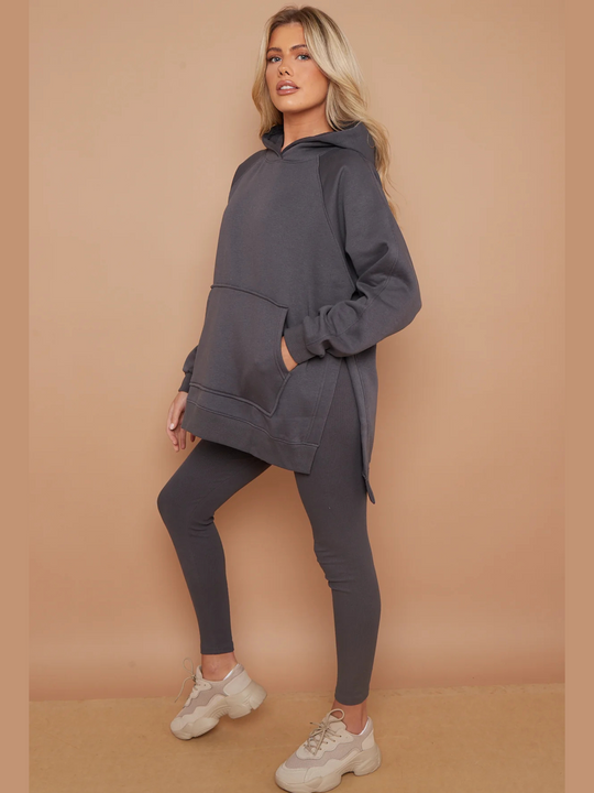 Full length shot. Model wears a charcoal grey oversized hoodie with front functional pockets and matching  grey ribbed leggings. The oversized hoodie has side splits on either side of the jumper.  Model has blonde hair,  she stands to the side. The full length of her body can be seen, demonstrating the side splits of the oversized hoodie. 
