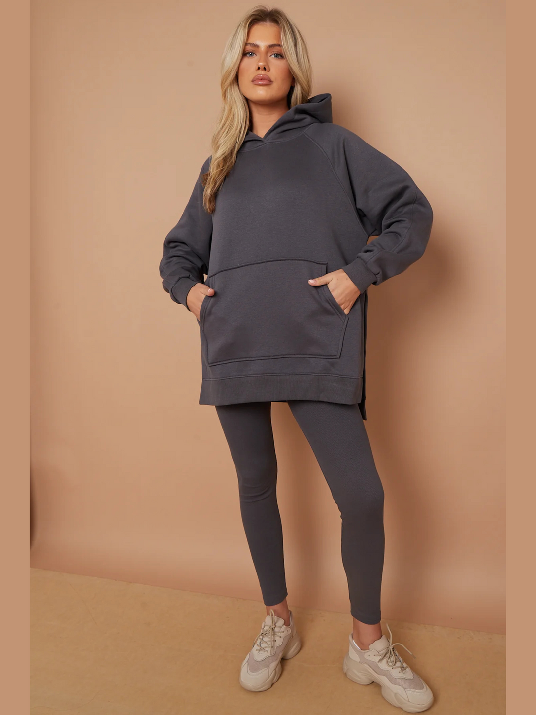 Full length shot. Model wears a charcoal grey oversized hoodie with front functional pockets and matching  grey ribbed leggings. The oversized hoodie has side splits on either side of the jumper.  Model has blonde hair,   she looks directly into the camera. She places both hands in the front functional pockets and places one leg slightly to the side. Model wears cream trainers  on her feet. 