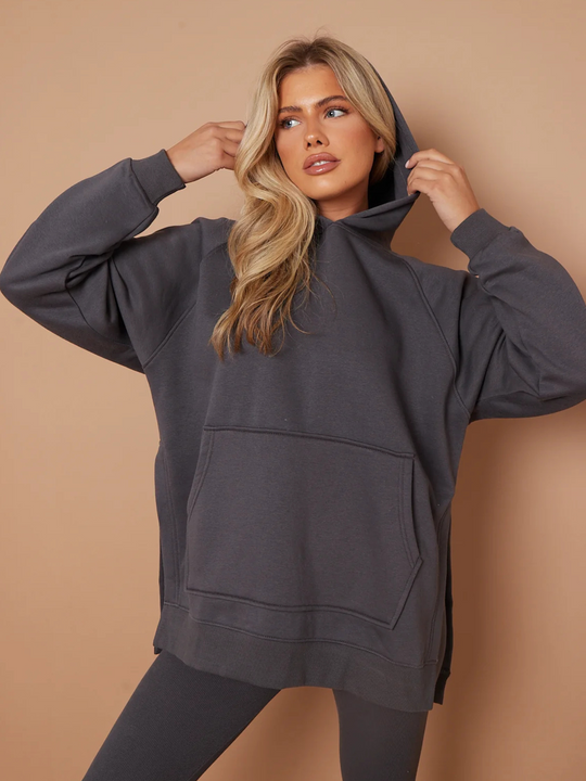Model wears a charcoal grey oversized hoodie with front functional pockets and matching  grey ribbed leggings. The oversized hoodie has side splits on either side of the jumper.  Model has blonde hair, looks to the side and places both hands on either side of the hoodie that is on her head. She places one leg slightly to the side, the ribbed leggings can be seen. 