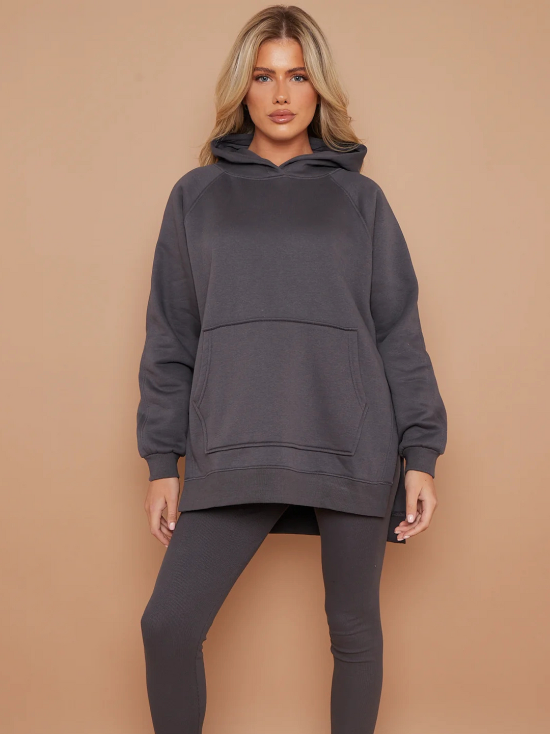 Model wears a charcoal grey oversized hoodie with front functional pockets and matching  grey ribbed leggings. The oversized hoodie has side splits on either side of the jumper.  Model has blonde hair,  she stands facing the camera placing both hands by her sides. 
