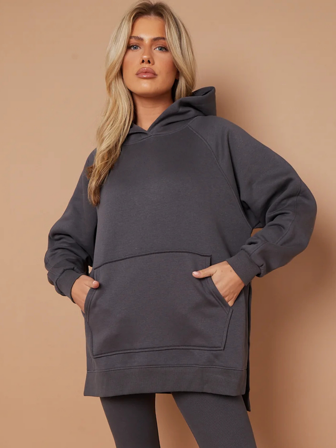 Model wears a charcoal grey oversized hoodie with front functional pockets and matching  grey ribbed leggings. The oversized hoodie has side splits on either side of the jumper.  Model has blonde hair and faces the camera, placing her hands inside either side of the functional front pockets. She places one leg to the side, the matching ribbed leggings can be seen.