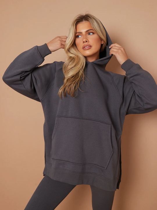 Model wears a charcoal grey oversized hoodie with front functional pockets and matching  grey ribbed leggings. The oversized hoodie has side splits on either side of the jumper.  Model has blonde hair, looks to the side and places both hands on either side of the hoodie that is on her head. She places one leg slightly to the side, the ribbed leggings can be seen. 