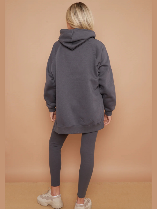 Full length shot. Model wears a charcoal grey oversized hoodie with front functional pockets and matching  grey ribbed leggings. The oversized hoodie has side splits on either side of the jumper.  Model has blonde hair,  she stands with her back to the camera; the back of the matching co-ord set can be seen including the hood of the hoodie and the  long length of the matching oversized jumper and ribbed leggings. 