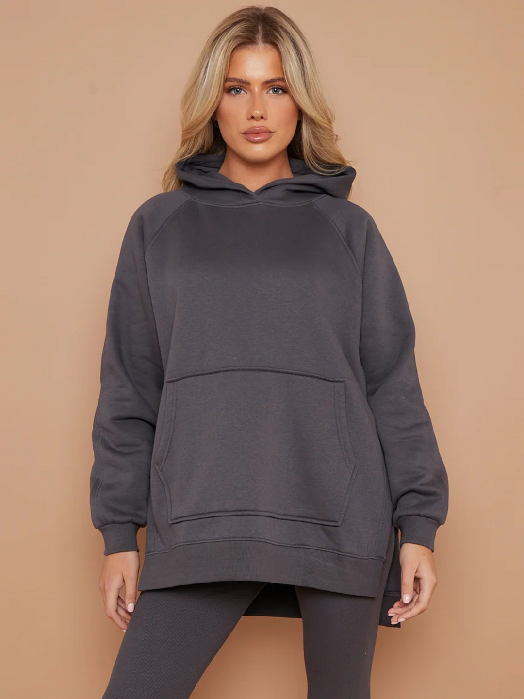 Model wears a charcoal grey oversized hoodie with front functional pockets and matching  grey ribbed leggings. The oversized hoodie has side splits on either side of the jumper.  Model has blonde hair,  she stands facing the camera placing both hands by her sides. 