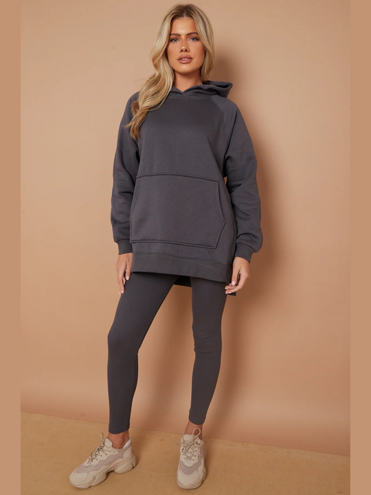 Full length shot. Model wears a charcoal grey oversized hoodie with front functional pockets and matching  grey ribbed leggings. The oversized hoodie has side splits on either side of the jumper.  Model has blonde hair,  she places both hands by her  sides and looks directly into the camera. Model wears cream trainers. 