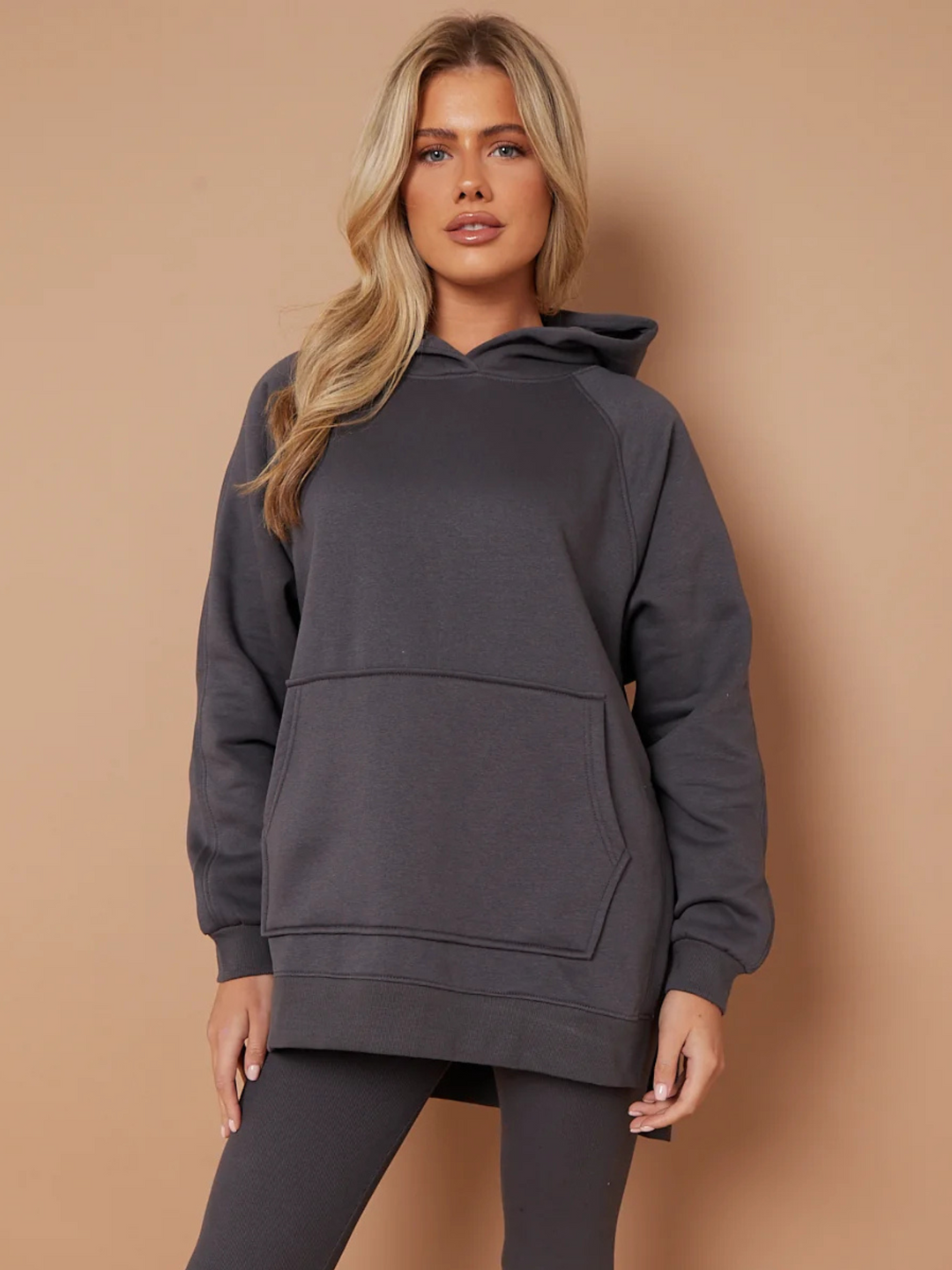 Model wears a charcoal grey oversized hoodie with front functional pockets and matching  grey ribbed leggings. The oversized hoodie has side splits on either side of the jumper.  Model has blonde hair,  she stares directly into the camera and places both hands by her sides. 
