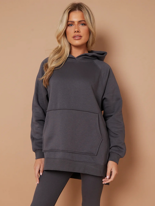 Model wears a charcoal grey oversized hoodie with front functional pockets and matching  grey ribbed leggings. The oversized hoodie has side splits on either side of the jumper.  Model has blonde hair,  she stares directly into the camera and places both hands by her sides. 