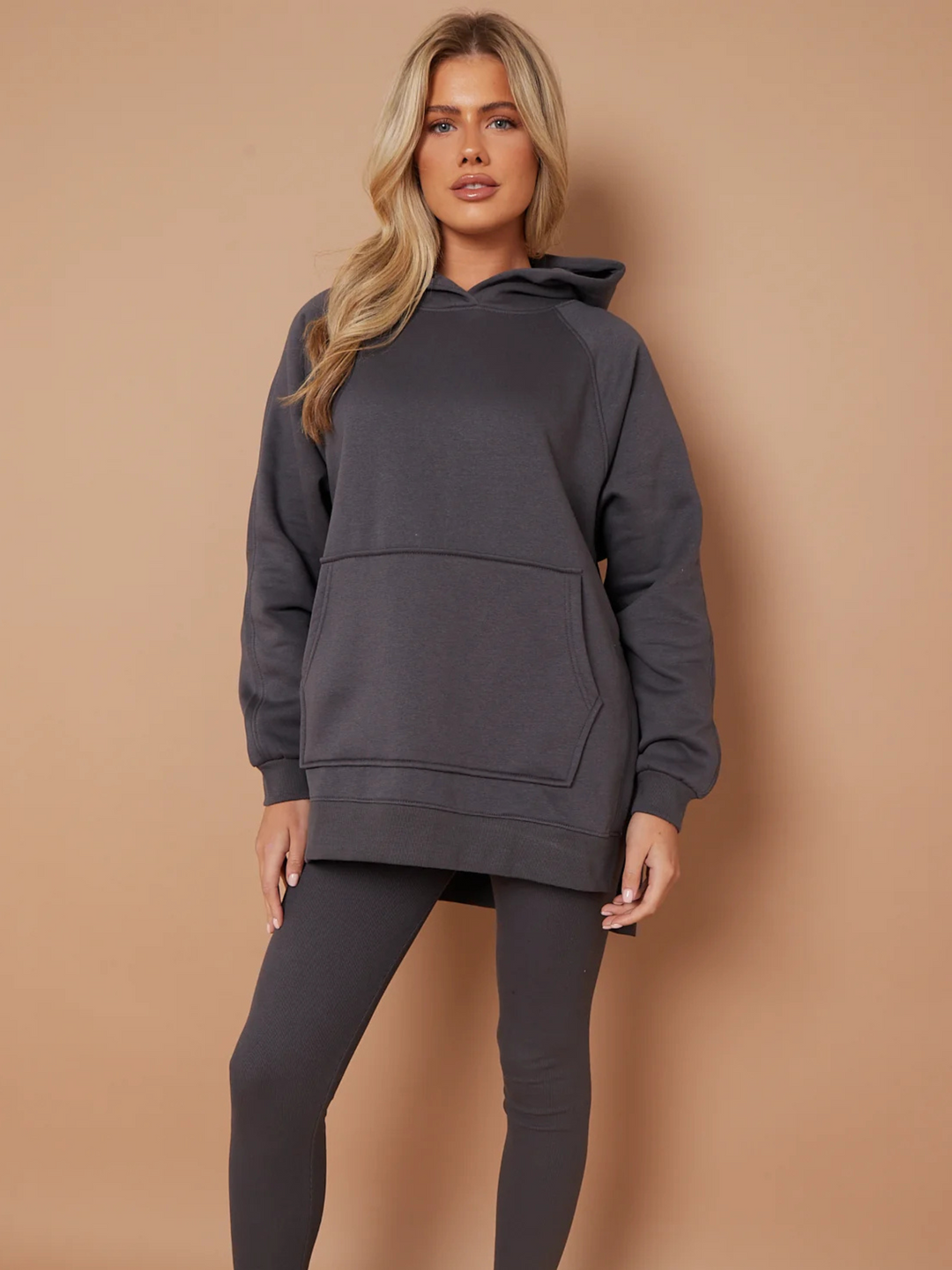 Model wears a charcoal grey oversized hoodie with front functional pockets and matching  grey ribbed leggings. The oversized hoodie has side splits on either side of the jumper.  Model has blonde hair,  she stands facing the camera, placing both hands bu her sides and placing one leg slightly to the side. 