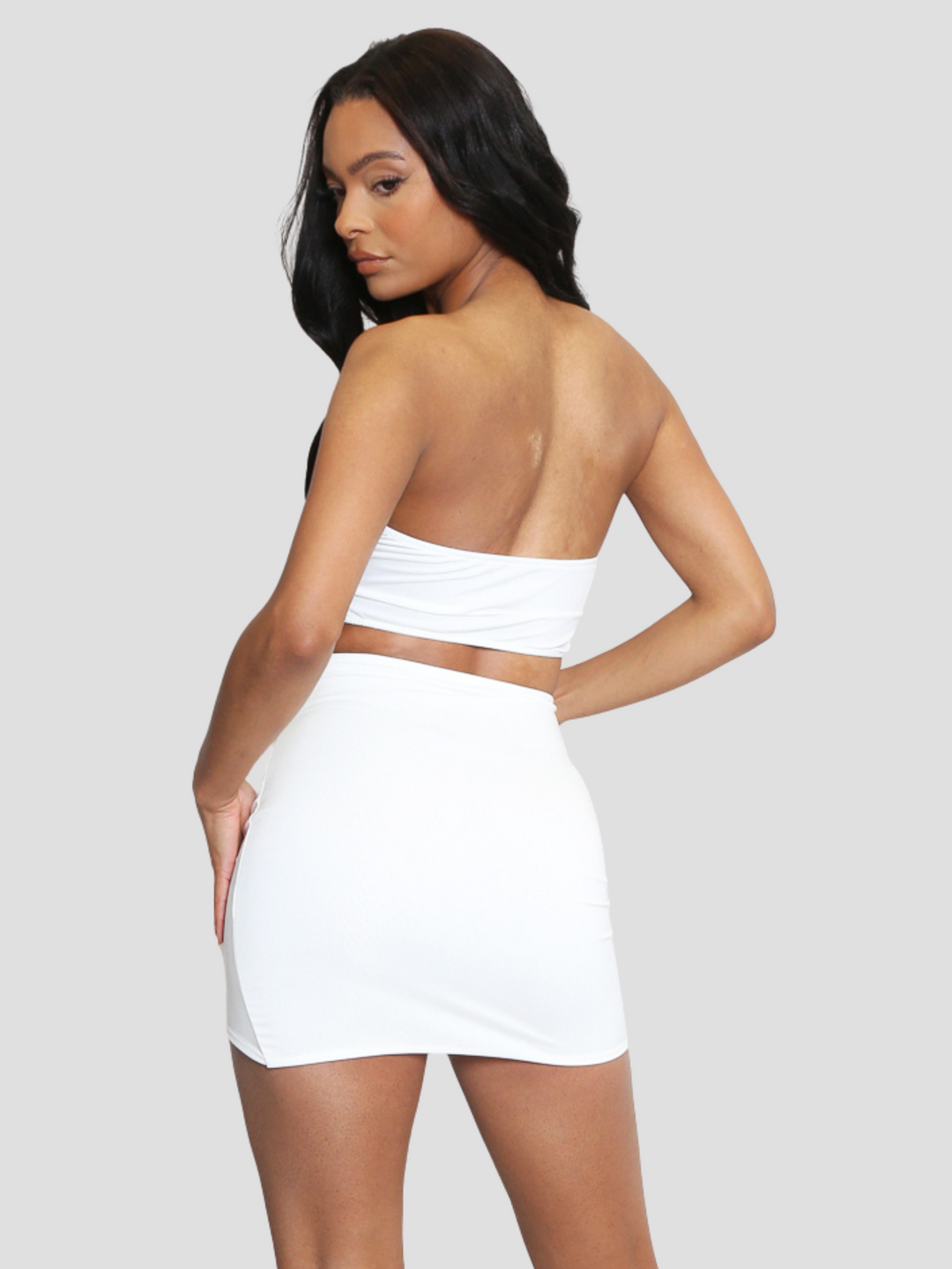 Female model stands in front of a plain light taupe background. The model wears a cream bandeau crop top with a corsage at the centre, she wears a matching cream high waisted mini skirt that also has a corasge detailing. The model has light brown skin and long black hair. She stands with her back to the camera, te back of the crop top and skirt can be seen.