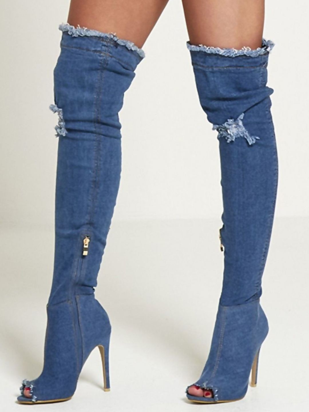 Model wears a dark denim thigh high boot that has a peep toe design and freyed edges and a distressed denim look. The boots has a stiletto heel and side zip fasteinf. The distressed denim boot has an over the knee desgn. The model stands to the side, one foot shows  the front of the boot and the other the inside of the boot, including zipped fastening.