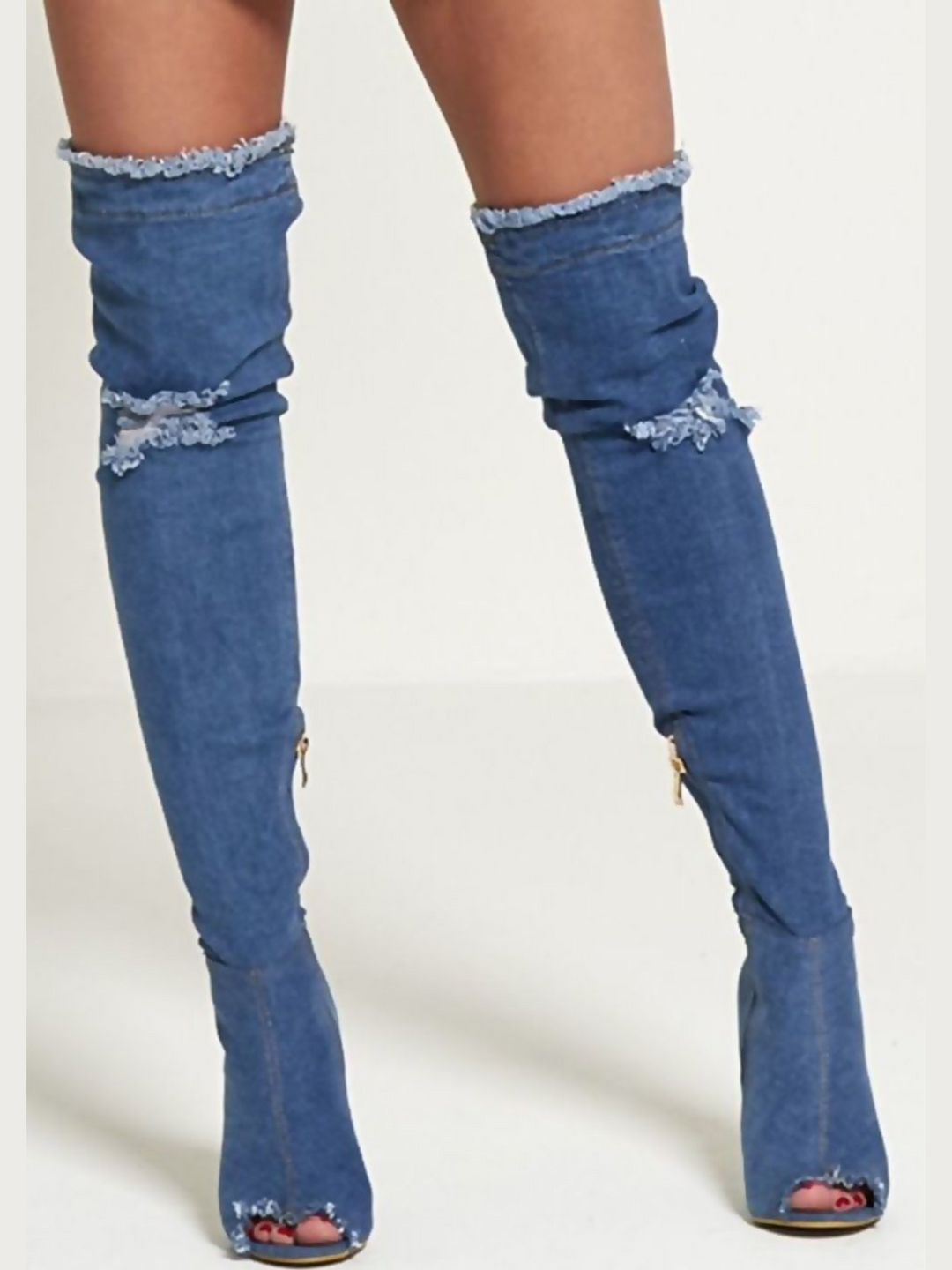 Model wears a dark denim thigh high boot that has a peep toe design and freyed edges and a distressed denim look. The boots has a stiletto heel and side zip fasteinf. The distressed denim boot has an over the knee desgn. The model faces forward, with both feet apart. The peep toe front of the boot can be seen. 