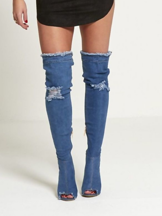 Model wears a dark denim thigh high boot that has a peep toe design and freyed edges and a distressed denim look. The boots has a stiletto heel and side zip fasteinf. The distressed denim boot has an over the knee design. The model faces forward, the front of the boots can be seen, including the peep toe design. 