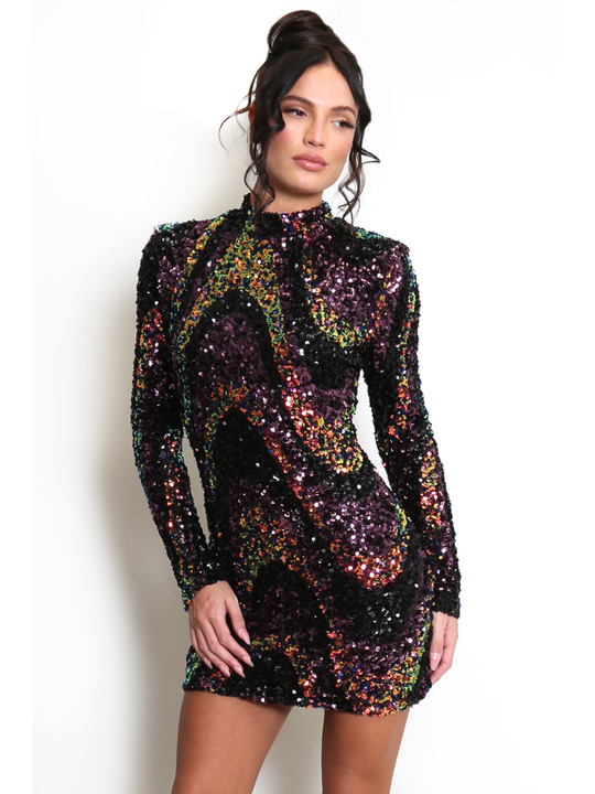 Female stands in front of a light cream wall. The model wears a long sleeve multicoloured sequin mini dress, with a mock neck. The model  has tanned skin and brown hair, she faces the camera and looks off to the side she places one hand by her side and the other on her thigh.