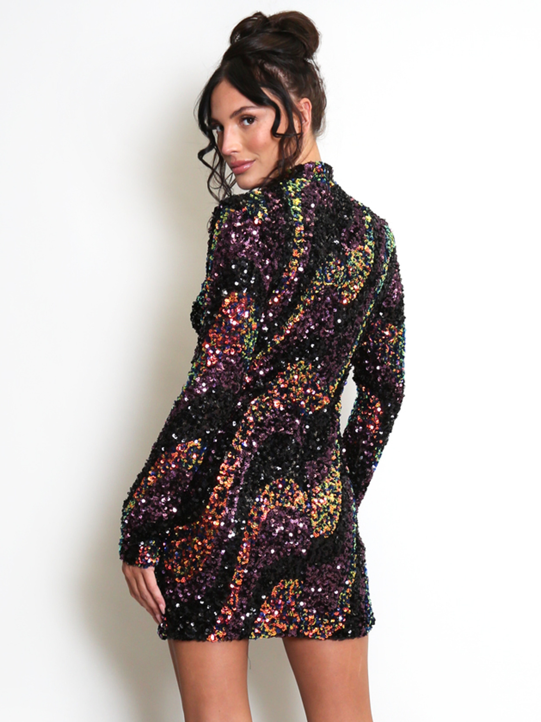Female stands in front of a light cream wall. The model wears a long sleeve multicoloured sequin mini dress, with a mock neck. The model  has tanned skin and brown hair, she stands with her back to the camera and turns her head to look into the camera. The back of the multicoloured sequin dress can be seen. 