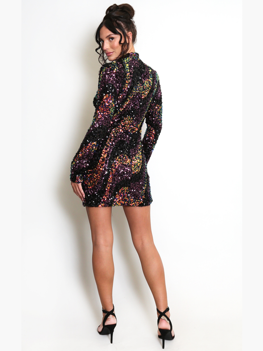 Full length shot. Female stands in front of a light cream wall. The model wears a long sleeve multicoloured sequin mini dress, with a mock neck. The model has tanned skin and brown hair, she stands with her back to the camera and turns her head to look into the camera, she wears black stiletto heels. 