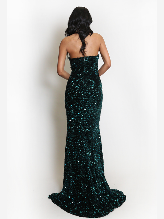 Full length shot. Model stands before a plain background. The model wears a green sequin maxi dress, with a fish tail hem and silver sequin sweetheart neckline. The model stands with her back to the camera, she has light brown skin and long black hair. The back of the maxi dress can be seen, including the zip back fastening and green luxe fish tail.