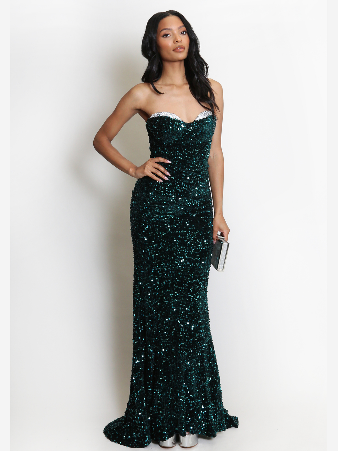 Full length shot. Model stands before a plain background. The model wears a green sequin maxi dress, with a fish tail hem and silver sequin sweetheart neckline. The model faces the camera, she has light brown skin and long black hair. She stands with one hand on her hip and the other by her side. The green sequin fish tail can be seen.