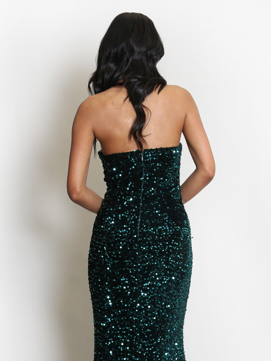 Full length shot. Model stands before a plain background. The model wears a green sequin maxi dress, with a fish tail hem and silver sequin sweetheart neckline. The model has light brown skin and long black hair, she stands with her back tl the camera. The  back of the dress can be seen, including the  concealed zip back fastening. 