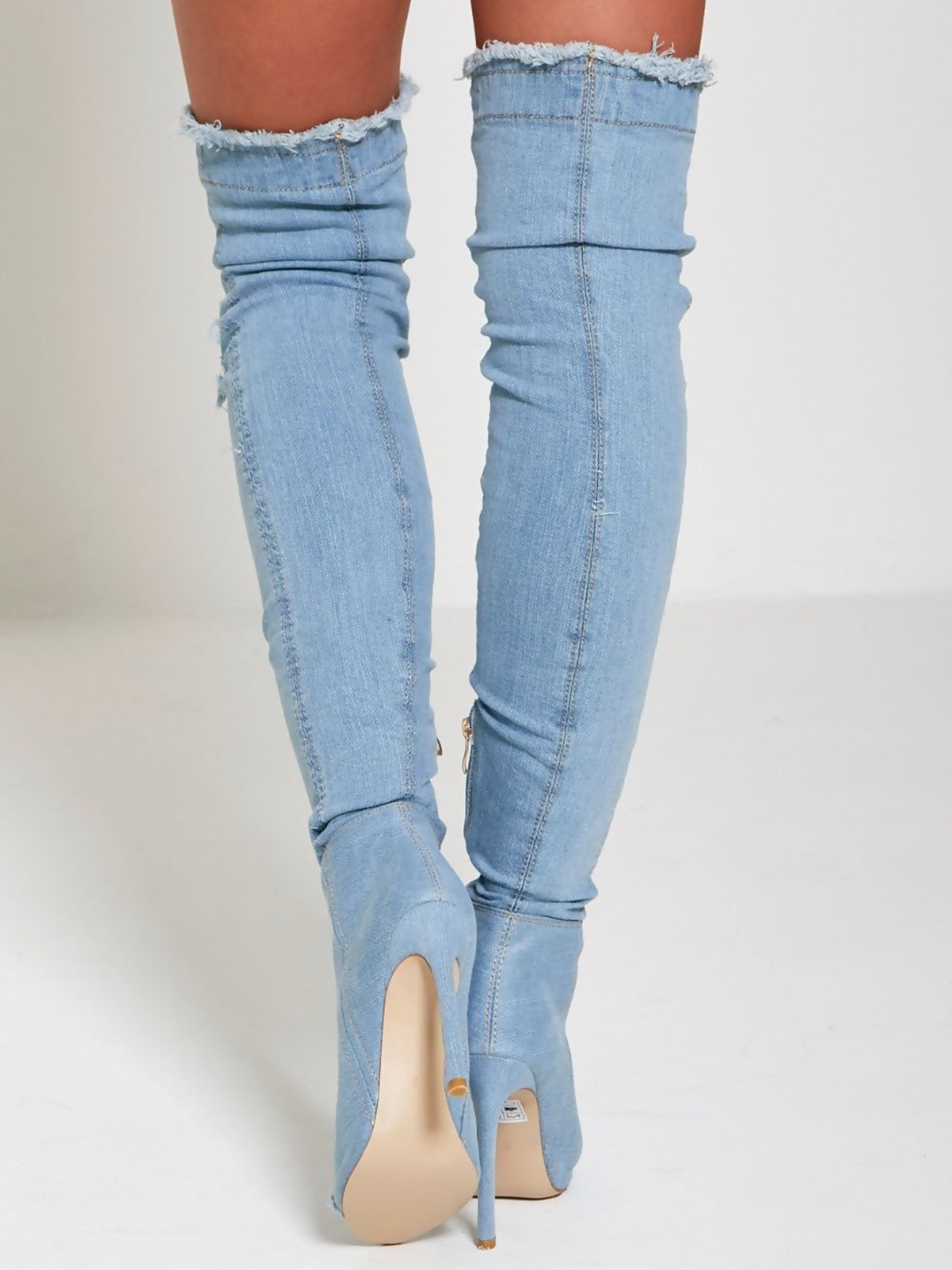 Model wears a light denim high heeled boots that has a distressed denim look. This over the knee thigh high boot has a peep toe design and a stiletto heel. Model stands with her back to the camera, the back of the boots can be seen, including thhe freyed distressed denim edges and the stiletto shoe. 