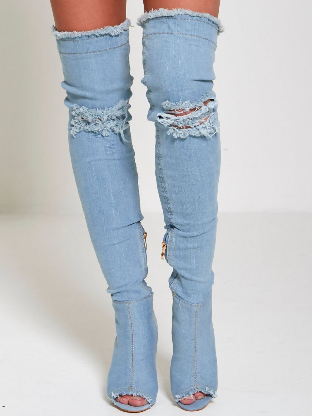 Model wears a light denim high heeled boots that has a distressed denim look. This over the knee thigh high boot has a peep toe design and a stiletto heel. The freyed denim at the edges of the thigh high boot and the peep toe is visbile. The freyed distressed design is also visible at the knee. 