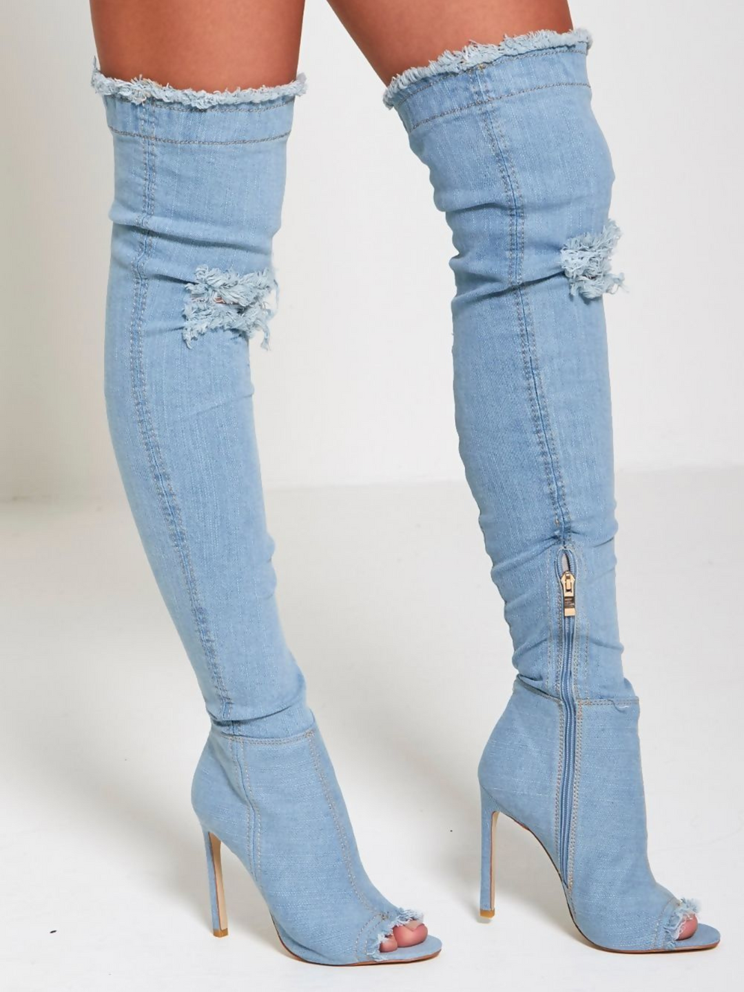 Model wears a light denim high heeled boots that has a distressed denim look. This over the knee thigh high boot has a peep toe design and a stiletto heel. The model stands to the side and both boots can be seen. The inside zip can be see on one of the boots and the front of the boot can be seen on the other. 