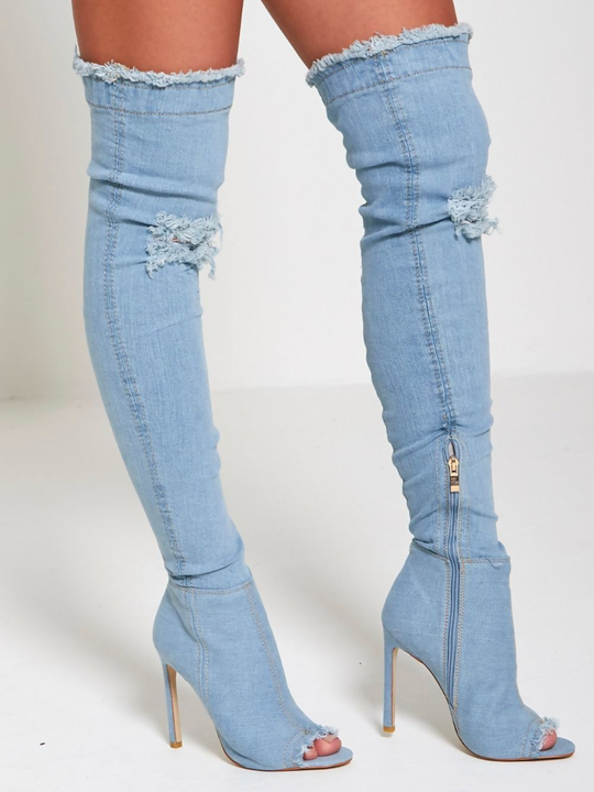 Model wears a light denim high heeled boots that has a distressed denim look. This over the knee thigh high boot has a peep toe design and a stiletto heel. The model stands to the side and both boots can be seen. The inside zip can be see on one of the boots and the front of the boot can be seen on the other. 
