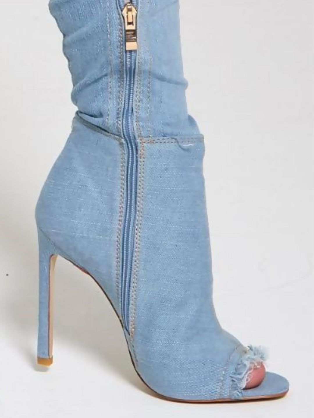Close up. Model wears a light denim high heeled boots that has a distressed denim look. This over the knee thigh high boot has a peep toe design and a stiletto heel. The bottom of the shoe is visible, the peep toe and the inside zip fastening is visible, including the freyed peep toe edges. 