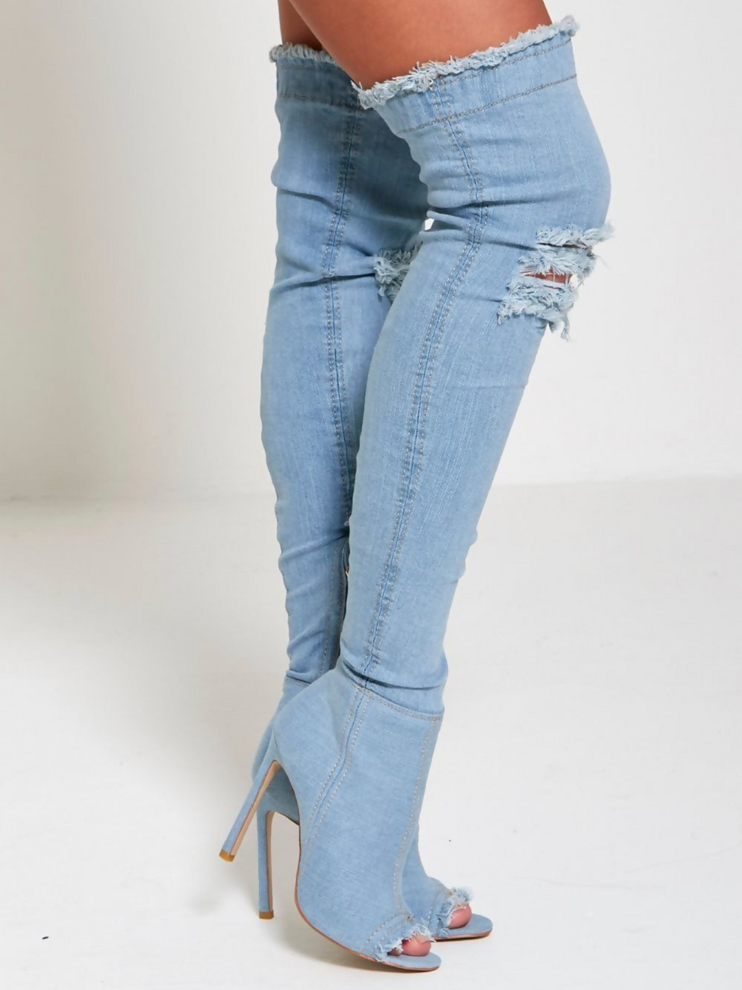 Model wears a light denim high heeled boots that has a distressed denim look. This over the knee thigh high boot has a peep toe design and a stiletto heel.  Model stands to the side, the peep toe and distressed denim an be seen. 