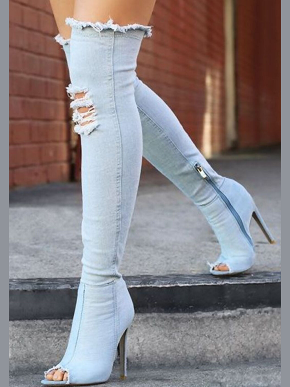Model wears a light denim high heeled boots that has a distressed denim look. This over the knee thigh high boot has a peep toe design and a stiletto heel. Model is outside and is walking down the steps both sides of the boots can be seen. 