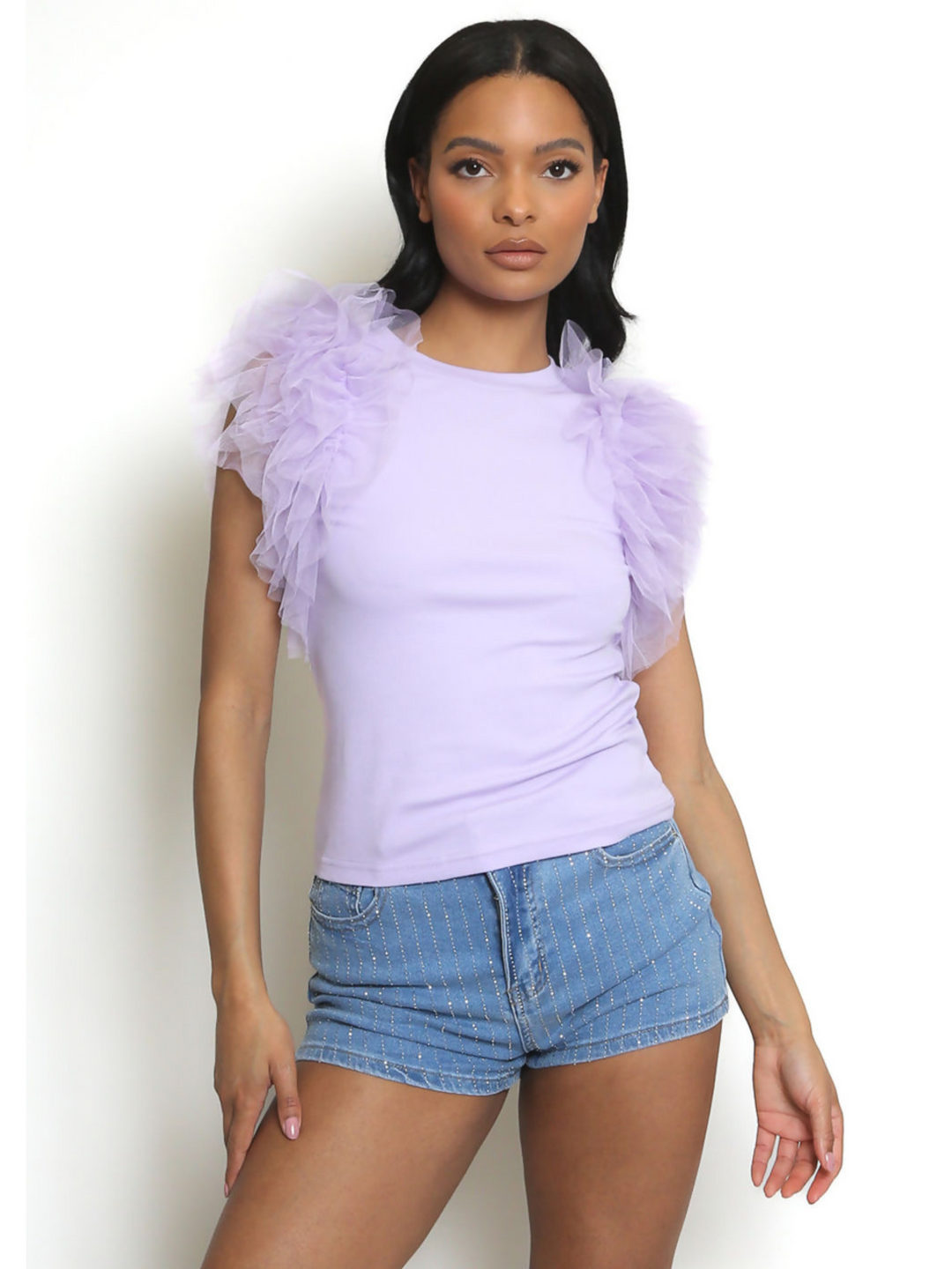 Woman wearing a light purple top with ruffled sleeves against a white background. The lilac t shirt has a rounded neck and the frill organza sleeves are short. The model has light brown skin and long black hair, she faces forward, looking directly into the camera with her hands by her sides, the front of the diamante shorts can be seen. 