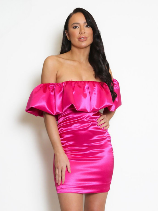 Model stands in front pf a plain background, she wears a fuchsia pink  satin mini dress with an off the shoulder frill design. The model has tanned skin and long black hair, she faces the camera and places one hand on her hip and the other on her thigh. Anne skin and long dark hair.
