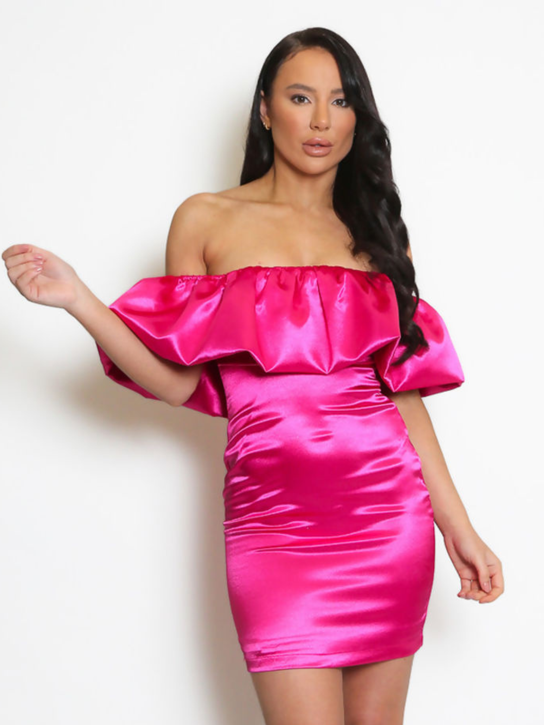 Model stands in front pf a plain background, she wears a fuchsia pink  satin mini dress with an off the shoulder frill design. The model has tanned skin and long black hair, she faces the camera, the front of the dress can be seen. 