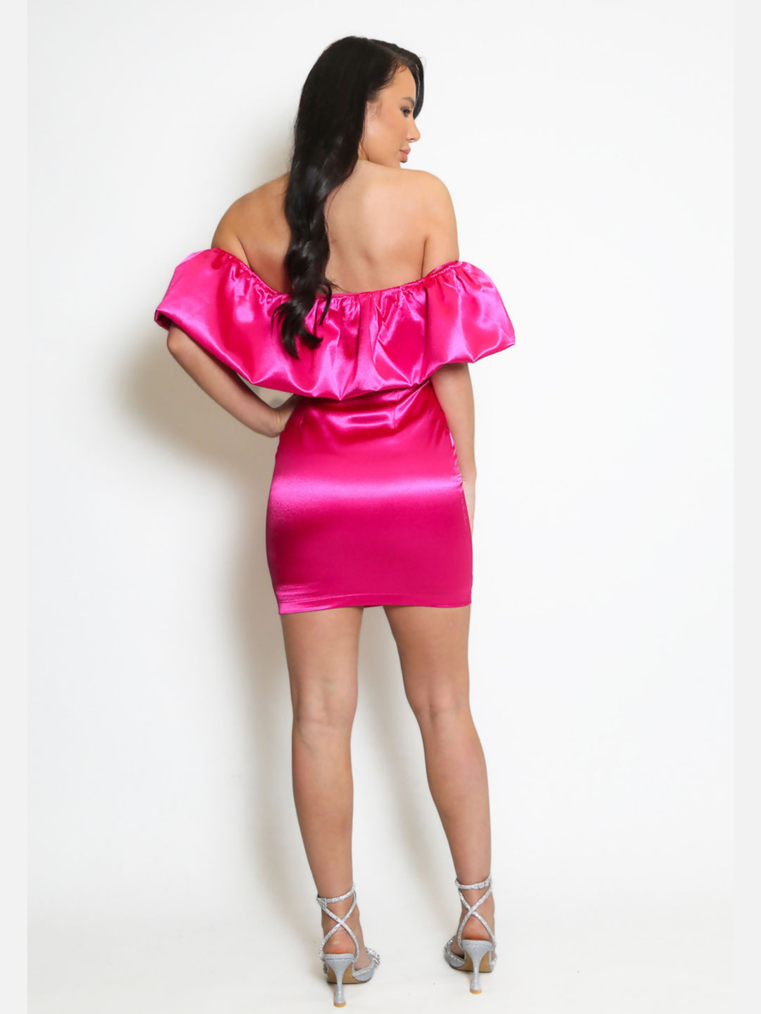 Model stands in front pf a plain background, she wears a fuchsia pink  satin mini dress with an off the shoulder frill design. The model has tanned skin and long black hair, she stands with her back to the camera, the back of the satin mini dress can be seen. 