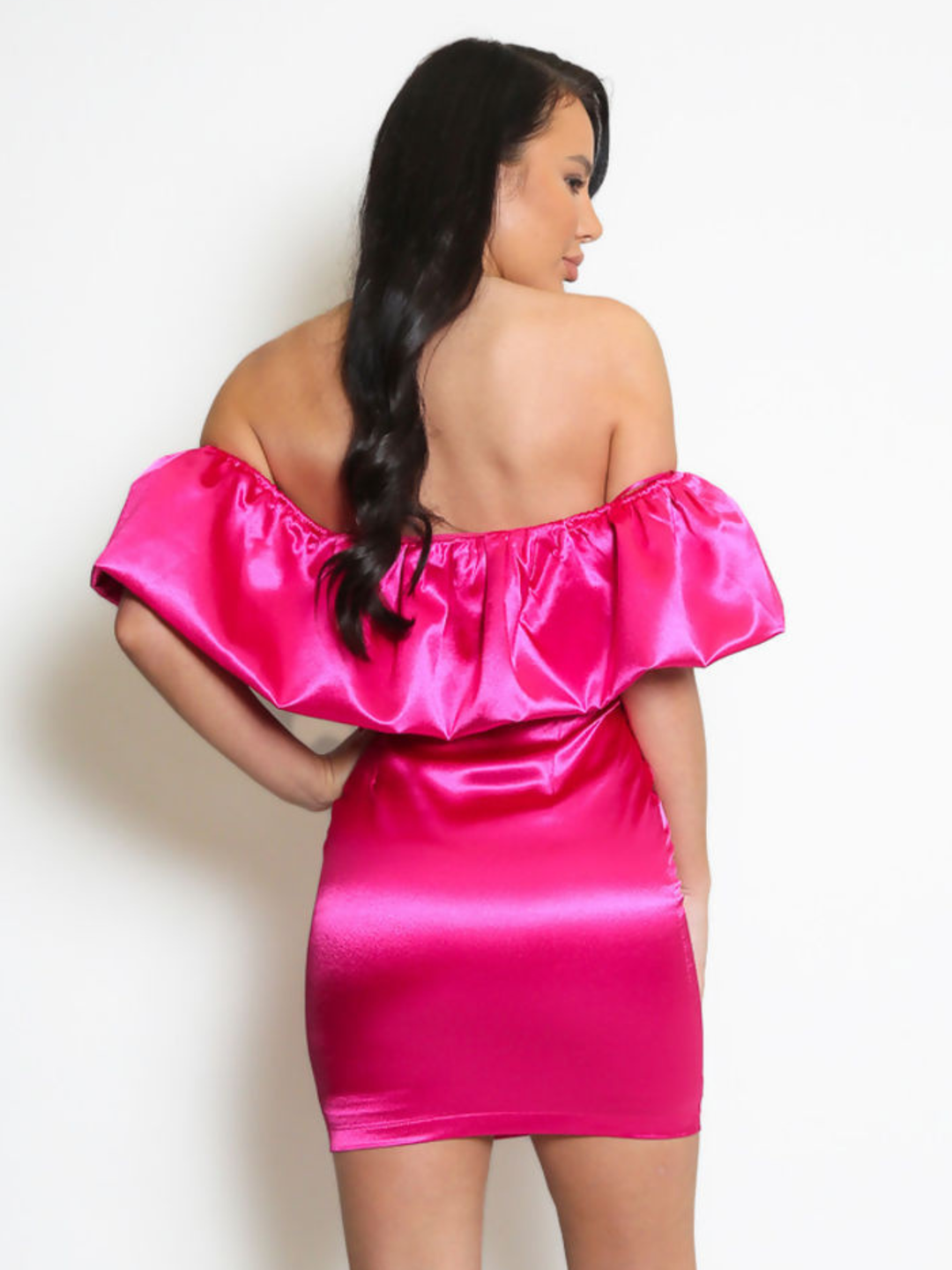 Model stands in front pf a plain background, she wears a fuchsia pink  satin mini dress with an off the shoulder frill design. The model has tanned skin and long black hair,  she stands with her back to the camera, placing one hand on her hip and the other by her side. The back of the satin dress can be seen. 