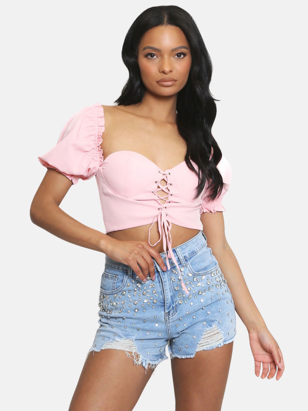 Model wears a pink sweet heart neckline and lace up front and puffy sleeves. Model has brown skin and black hair. Model faces the camera and places one hand on a diamante denim shorts and one arm by her side.