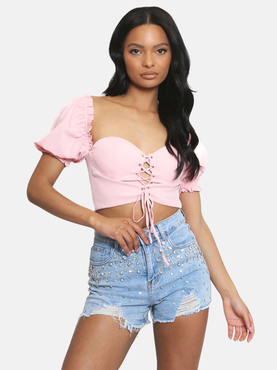 Model wears a pink sweet heart neckline and lace up front and puffy sleeves. Model has brown skin and black hair. Model faces the camera and places one hand on a diamante denim shorts and one arm by her side.
