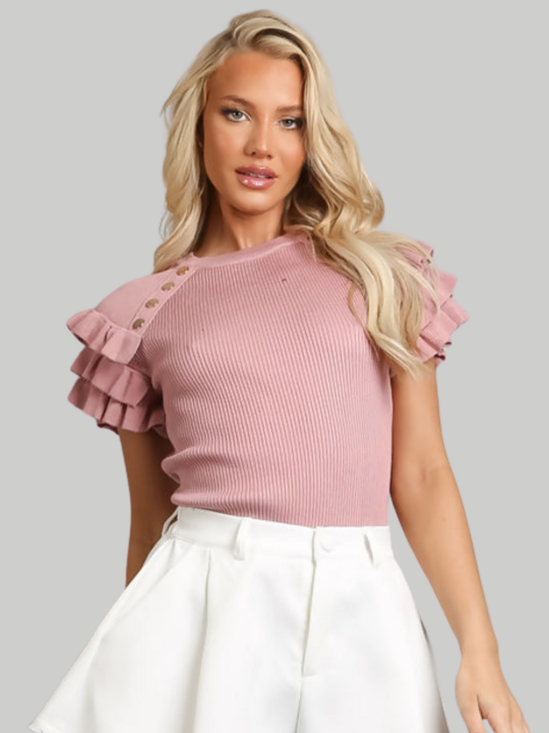 Female model wears a pink ribbed top with ruffled 3/4 length top and and gold buttons detailing and wears white pants on a gray background. The model has tanned skin and blonde hair, she faces forward with one hand by her side and the other behind her back. 