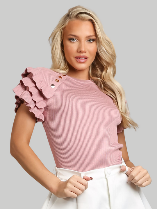 Female model wears a pink ribbed top with ruffled 3/4 length top and and gold buttons detailing and wears white pants on a gray background. The model has tanned skin and blonde hair,  she faces the camera and places her hands in the belted waist of the shorts. 