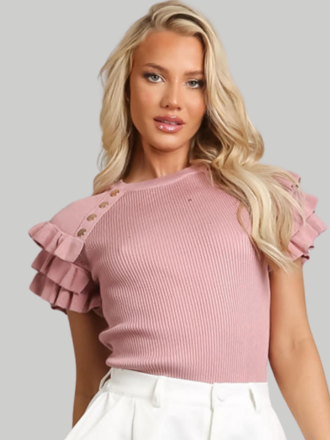 Female model wears a pink ribbed top with ruffled 3/4 length top and and gold buttons detailing and wears white pants on a gray background. The model has tanned skin and blonde hair, she looks directly into the camera and places one hand behind her back and the other by her side. 