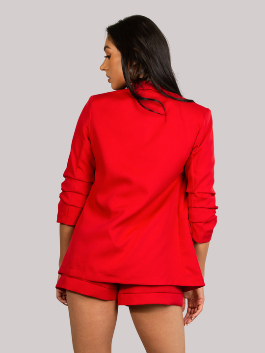 Female model stands in front of a plain background. The model wears a red open blazer with ruched 3/4 sleeves and a matching red shorts. The  model has tanned skin and long black hair, she stands with her back to the camera; the back of the red blazer can be seen, including the ruched sleeves. The back of the red short is also visible. 