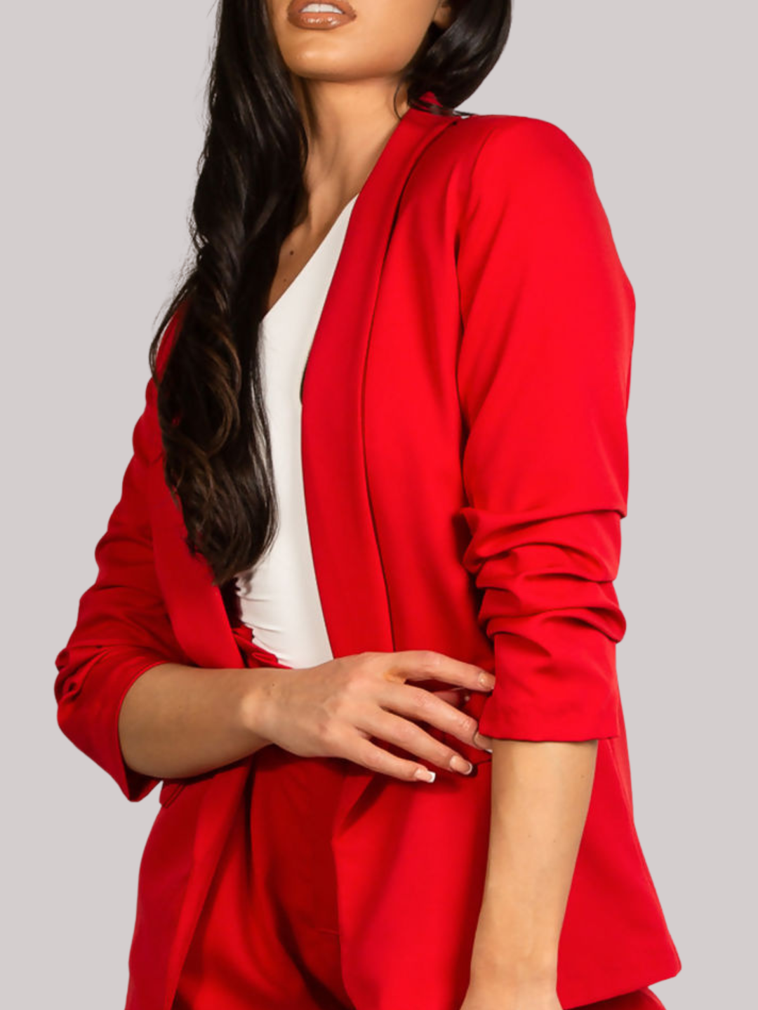 Close up. Female model stands in front of a plain background. The model wears a red open blazer with ruched 3/4 sleeves and a matching red shorts. The  model has tanned skin and long black hair. The closeup shows the front of the blazer, including the 3/4 llength ruched sleeves. 