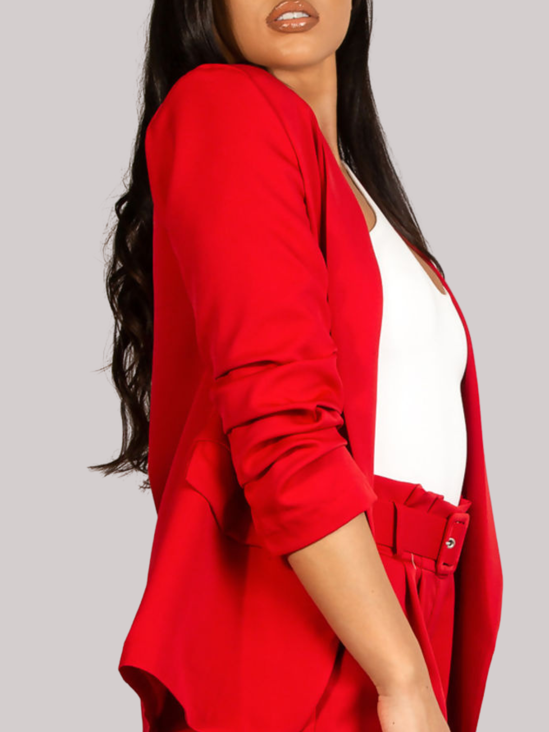 Close up. Female model stands in front of a plain background. The model wears a red open blazer with ruched 3/4 sleeves and a matching red shorts. The  model has tanned skin and long black hair, the close up of the garment shows the side of the red blazer and 3/4 length ruched sleeves, the red belted shorts can also be seen. 